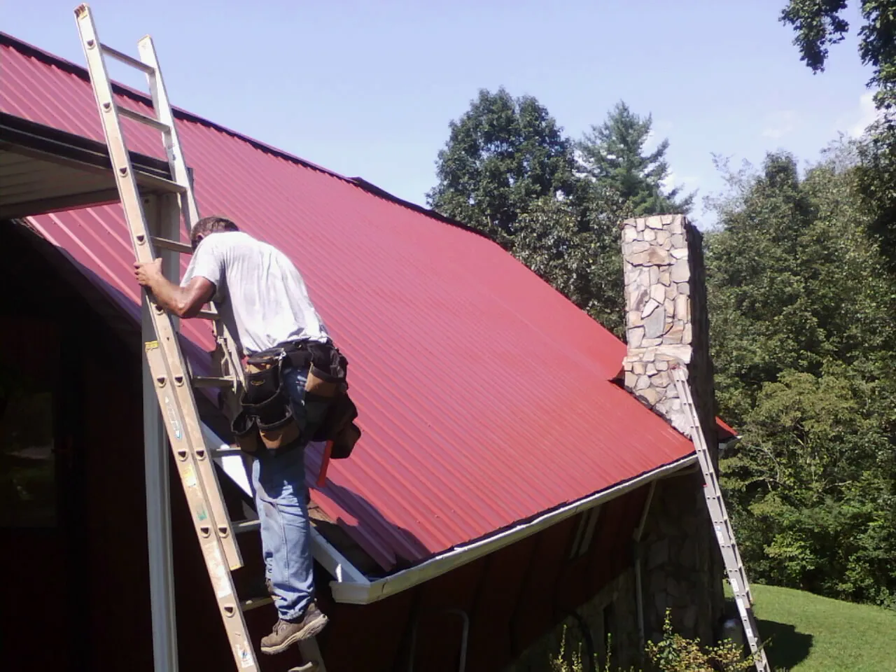 Roofer on a ladder beside a red metal roof and stone chimney — Soffit & Fascia Repair by Roofing Brothers in Moss Landing